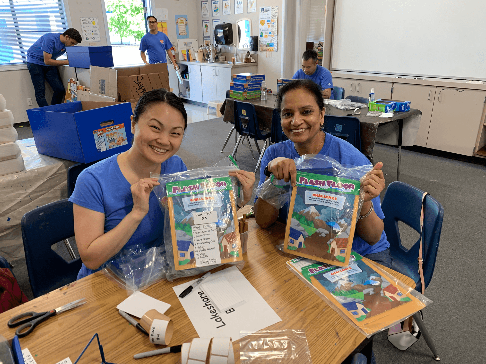 Two female employees of SIE volunteering to help communities prepare for a flash flood. The are holding up the flash flood kits and smiling.