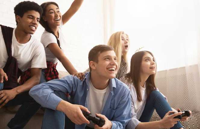 A diverse group of 5 young adults on a couch together watching two of the friends play a PS game as they are holding controllers and smiling.