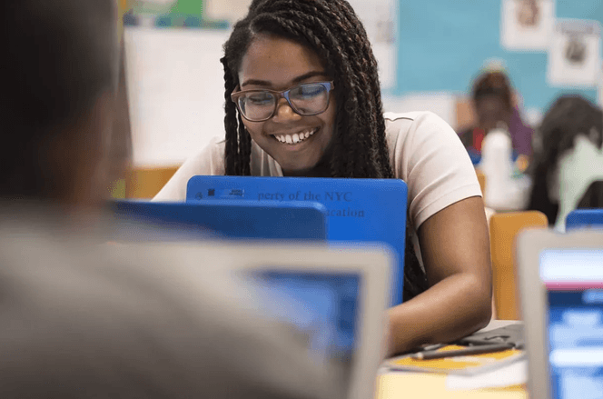 Young teenage girl with braids smiling at her laptop while coding in a classroom for GMG.