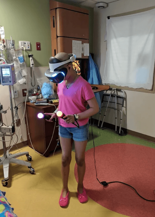 Young girl who's in a hospital being treated for cancer playing a VR game in her hospital room. She's dressed in pink with no hair.