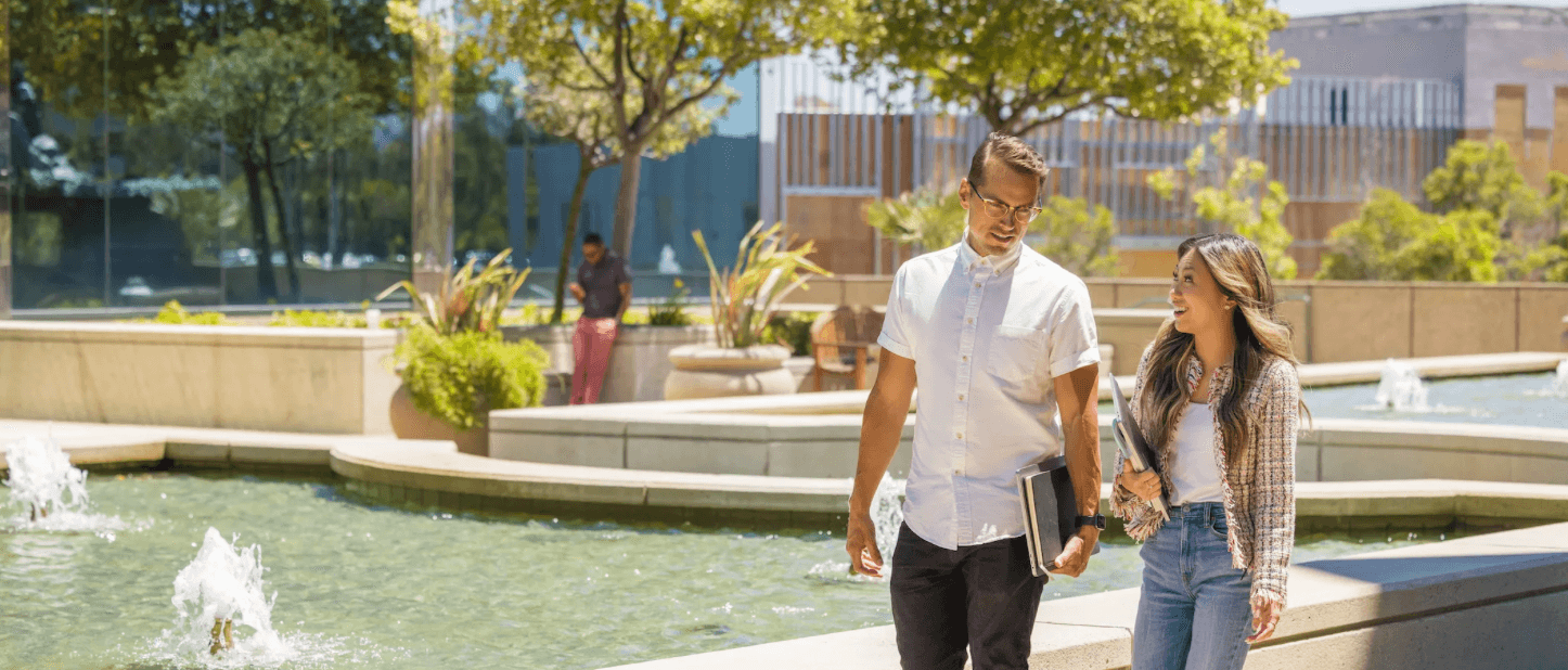 A man and woman walking through the outdoor campus of the San Mateo offices, there's fountains and greenery.