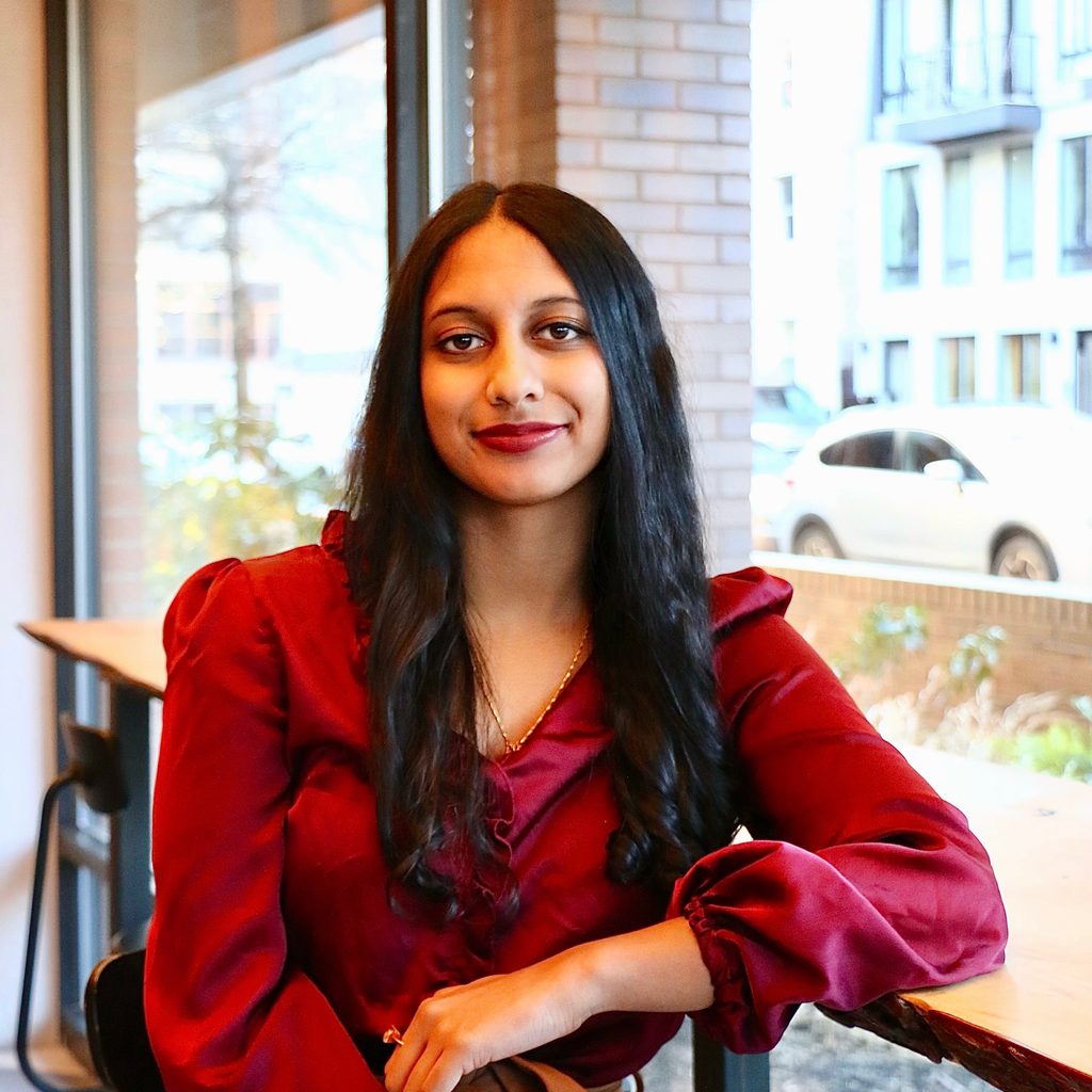 A headshot of a young woman with long dark hair and a red long sleeved shirt, sitting in a cafe.