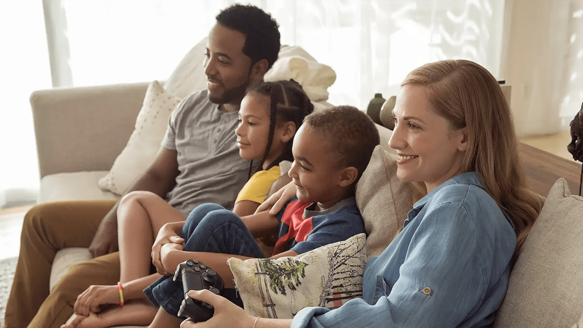 A mother holding a game controller plays video games alongside her husband, son, and daughter, all smiling together on a couch.