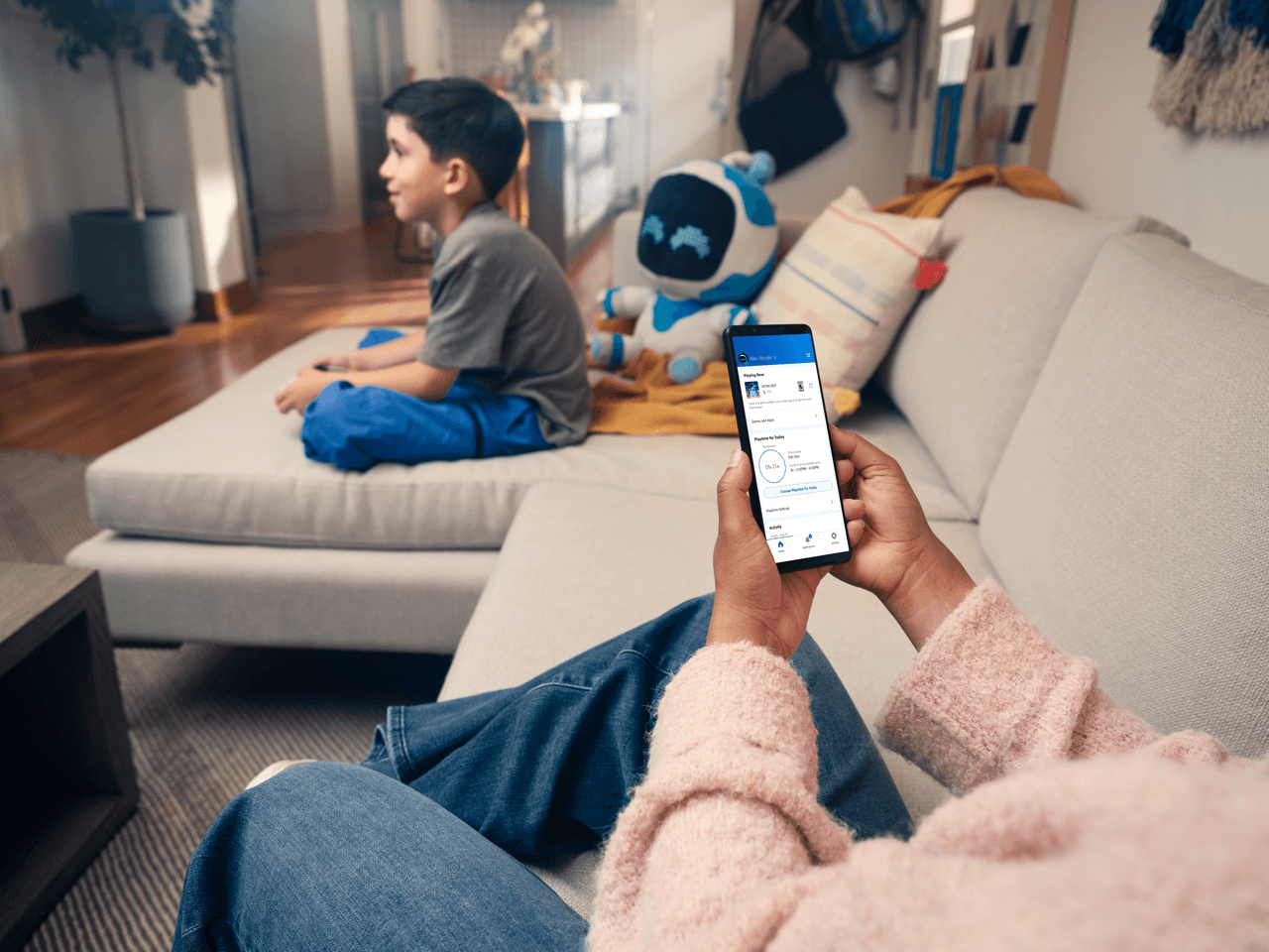 A parent is using the PlayStation Family app on their smartphone, while a boy plays games next to them on the couch.