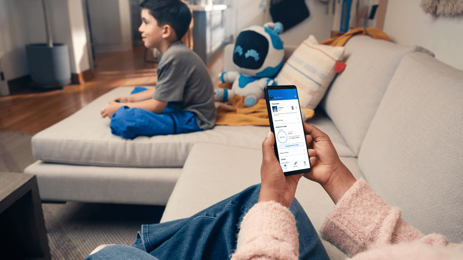 A parent is using the PlayStation Family app on their smartphone, while a boy plays games next to them on the couch.