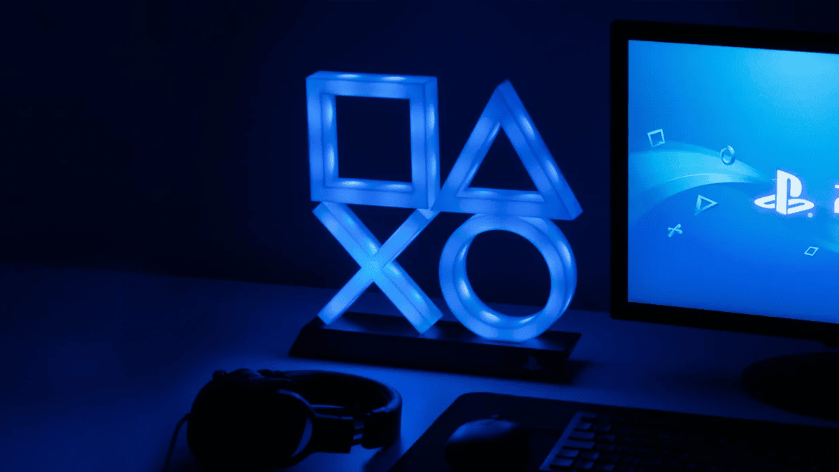 A light made up of PlayStation shapes (square, triangle, cross, circle) is glowing in a dark room, next to a monitor with a blue PlayStation background.