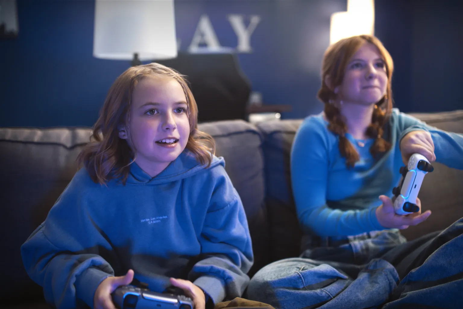 Two young girls in a blue hoodie sitting on a couch playing video games together with the PlayStation 5 DualSense wireless controller. 