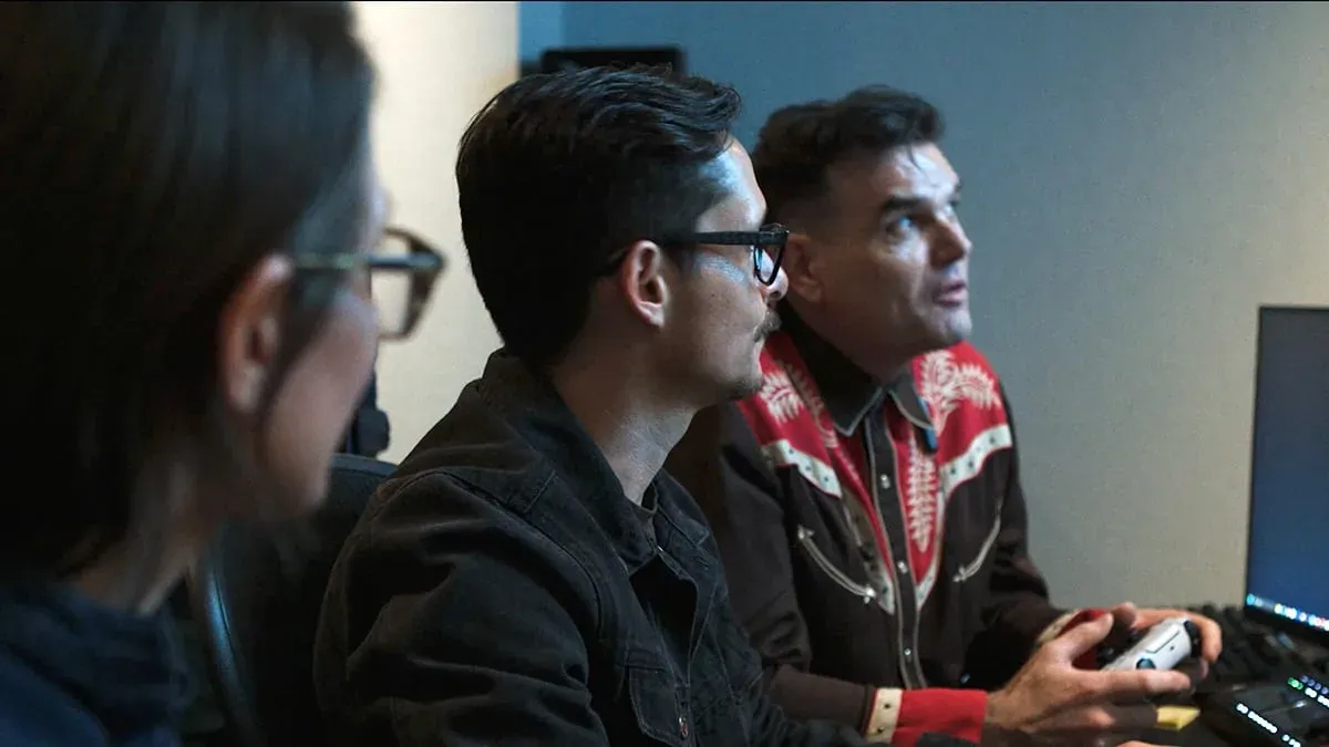 Behind-the-scenes studio photograph showing game developers reviewing project data. In the foreground, team members look at a monitor in profile, while Insomniac Games Creative Director Marcus Smith, wearing a distinctive red and black western shirt, holds a white gaming controller.