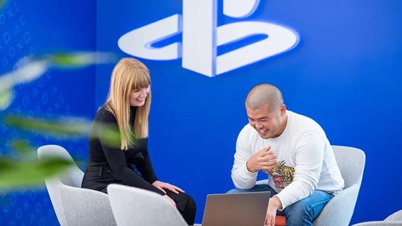 Two diverse employees smiling and collaborating over a laptop in front of a PlayStation logo wall.