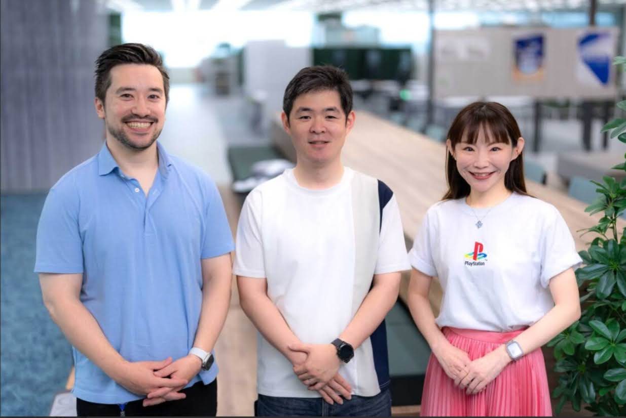 明るいオフィスで並んで立つ3名の社員が笑顔を向けている